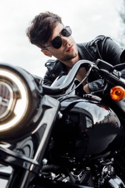 selective focus of handsome motorcyclist in leather jacket looking away while sitting on motorcycle clipart