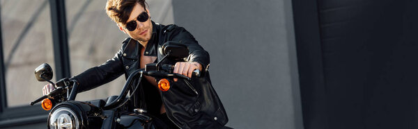 panoramic shot of motorcyclist in leather jacket sitting on motorcycle and looking away