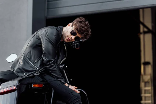 ガレージの近くにオートバイに座っている間 サングラスと革のジャケットを着た若いモーターサイクリスト — ストック写真