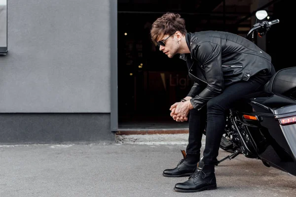 黒いオートバイに座って前方を見ている革のジャケットとサングラスを着たハンサムな男の側面図 — ストック写真