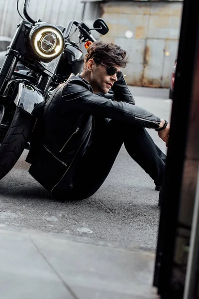 明るいランプを持つ黒いオートバイの近くに地面に座っているハンサムな若いモーターサイクリスト — ストック写真