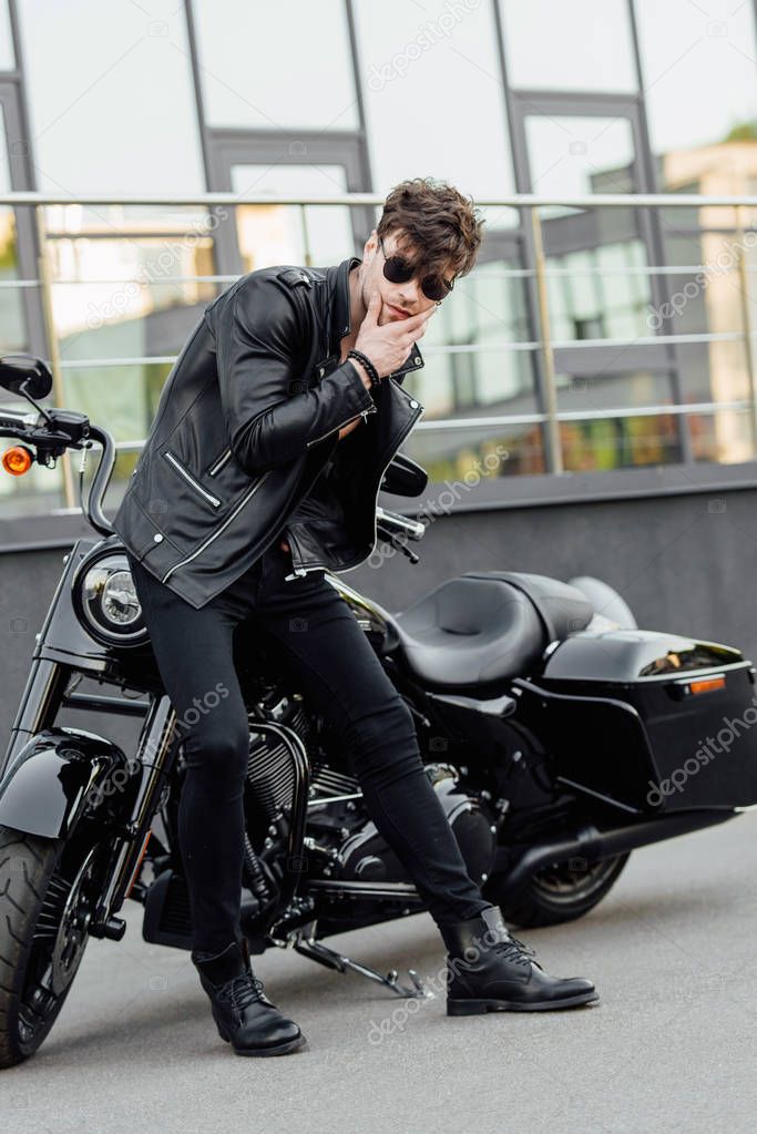 handsome young motorcyclist standing near motorcycle, looking away and touching chin