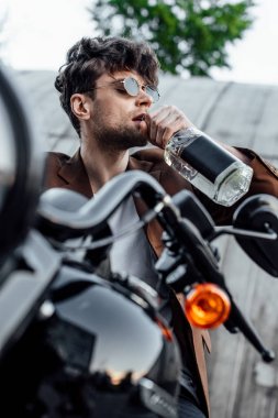 yakışıklı adam motosiklet üzerinde otururken alkol içme seçici odak ve uzağa bakıyor