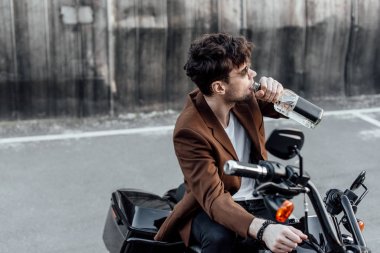 motosiklet üzerinde otururken şişeden alkol içen kahverengi ceketli adam
