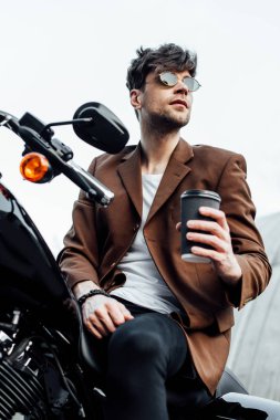motosiklet üzerinde otururken gitmek için kahve tutarak ceket ve güneş gözlüğü şık adam düşük açı görünümü
