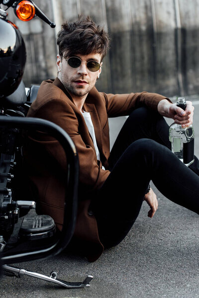 handsome man sitting near motorcycle, holding bottle with alcohol and looking away