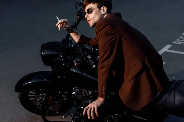 穿棕色夹克的年轻人坐在黑色摩托车上抽烟 — 图库照片