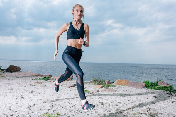 sportive blonde woman running and listening music in earphones near sea 