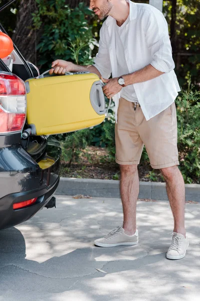 cropped view of happy man in cap putting travel bag in car trunk
