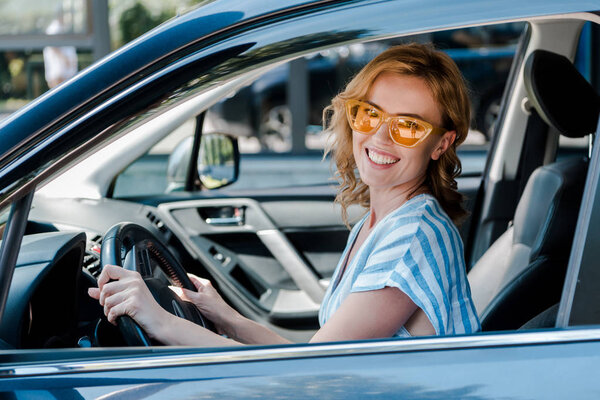 happy blonde woman in sunglasses holding steering wheel while sitting in car 