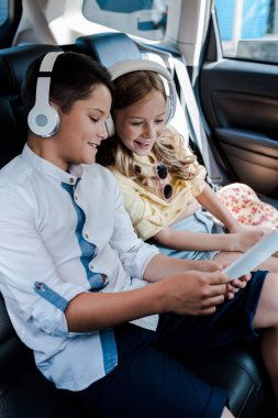 selective focus of cheerful kids listening music in headphones while looking at smartphone in car  clipart