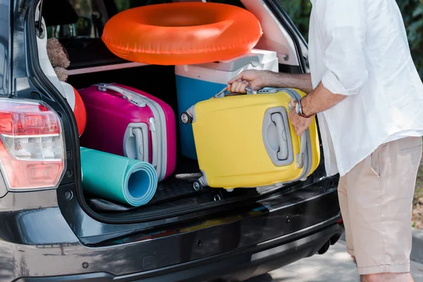 車のトランクにピンクのスーツケースの近くに黄色の荷物を置く男のトリミングビュー — ストック写真
