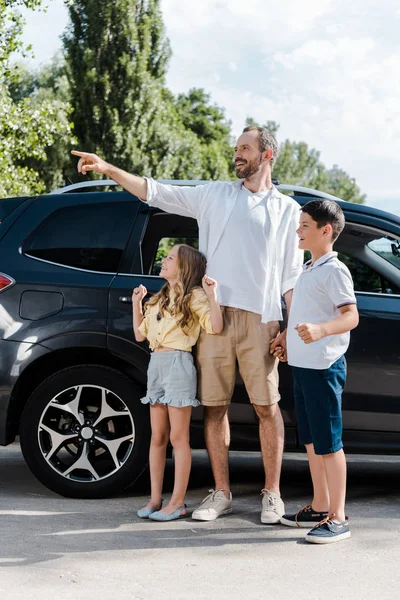 happy father pointing with finger while standing with cheerful kids and car