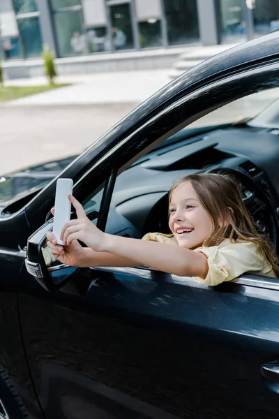 車の中に座っている間 スマートフォンで自分撮りを撮る幸せな子供の選択的な焦点 — ストック写真