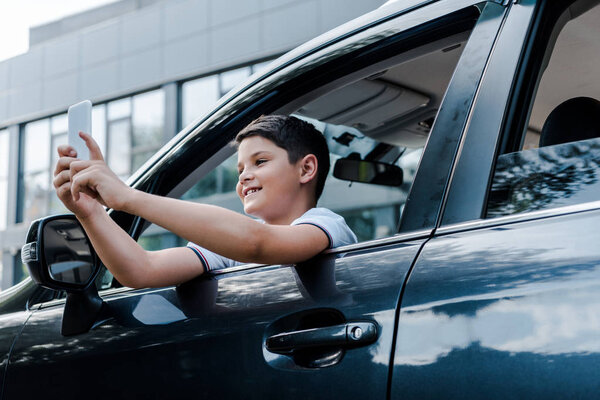 low angle view of happy kid taking selfie on smartphone in car near building 