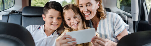 panoramic shot of cheerful family taking selfie on smartphone in car 