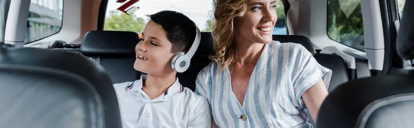 車の中で母の近くにヘッドフォンで陽気な子供のパノラマショット — ストック写真