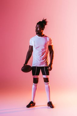 degrade ve aydınlatma ile pembe arka planda Rugby topu tutarak uzak görünümlü yakışıklı Afrikalı Amerikan sporcu