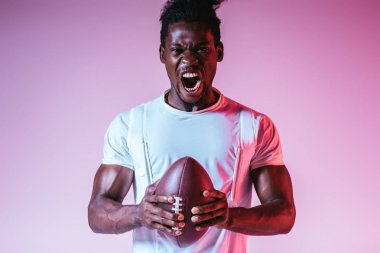 excited african american sportsman yelling at camera while holding rugby ball on purple background with gradient