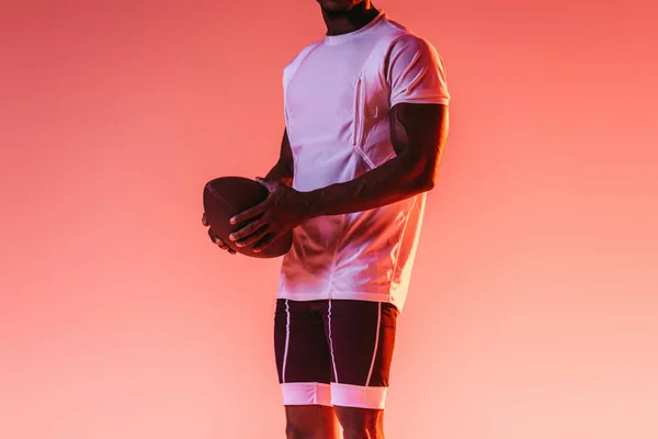 グラデーションと照明でピンクの背景にラグビーボールを保持するアフリカ系アメリカ人のスポーツマンの部分的なビュー — ストック写真