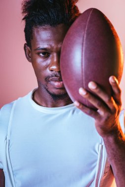 aydınlatma ile pembe arka planda Rugby topu tutarken ciddi Afrikalı Amerikan sporcu kamera bakıyor