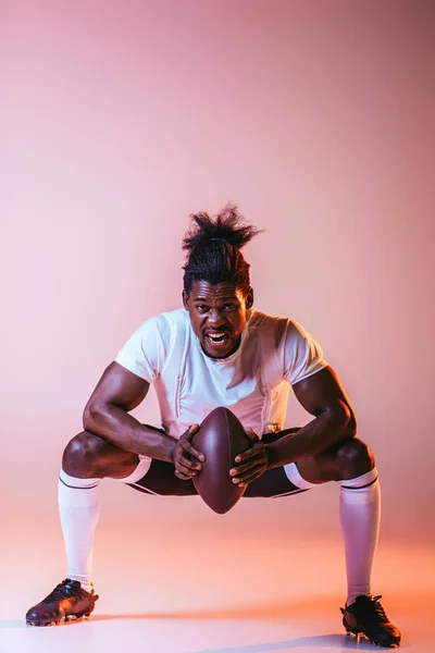 グラデーションと照明でピンクの背景にアメリカンフットボールをプレイしながら叫ぶ興奮アフリカ系アメリカ人スポーツマン — ストック写真