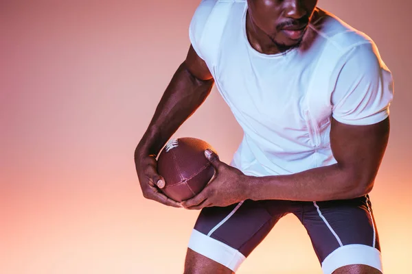 グラデーション付きピンクの背景にアメリカンフットボールをプレイするアフリカ系アメリカ人スポーツマンの部分的なビュー — ストック写真