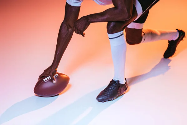 ピンクと黄色のグラデーションと照明を背景にアメリカンフットボールをプレイするアフリカ系アメリカ人スポーツマンのトリミングされたビュー — ストック写真
