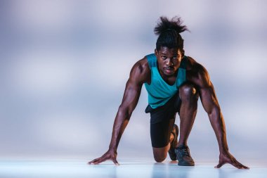 başlangıç pozisyonunda Afrika Amerikan sporcu ayakta ve aydınlatma ile gri arka planda kamera bakıyor