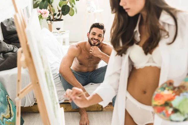 シャツのない男が微笑み ベッドに座っている間 白い下着の絵でセクシーな女の子のトリミングされたビュー — ストック写真