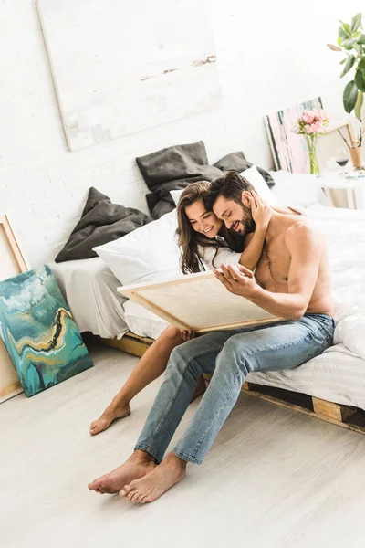 年轻夫妇坐在床上微笑 而男人拿着画和女孩拥抱的家伙 — 图库照片