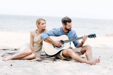 beautiful young barefoot woman sitting on blanket with boyfriend playing acoustic guitar at beach near sea clipart