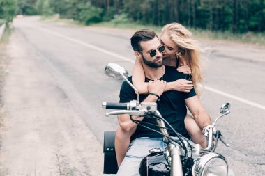 young tender couple of bikers hugging on black motorcycle on road near green forest clipart