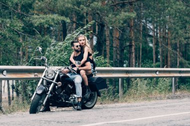 young couple of bikers hugging while sitting on black motorcycle on road near green forest clipart