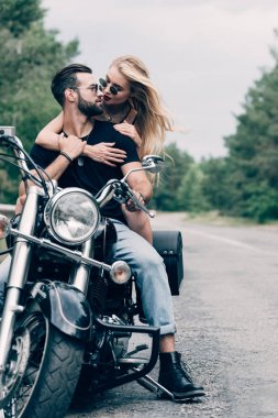 young couple of bikers closely looking at each other on black motorcycle on road near green forest clipart