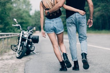 partial view of young couple of bikers walking along road and embracing near black motorcycle clipart