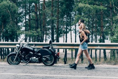 yol boyunca yürüyen ve siyah motosiklet ve yeşil orman yakınında kucaklayan bisikletçilerin genç çift yan görünümü