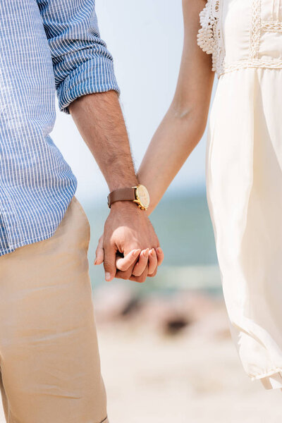 partial view of couple holding hands at beach in sunlight