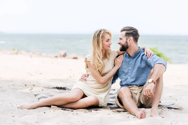 毛布の上に座って 海の近くのビーチでお互いを見て幸せな美しい若い裸足のカップル — ストック写真