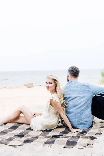 海の近くのビーチでアコースティックギターでボーイフレンドの近くにチェック柄の毛布の上に座って笑顔の若いブロンドの女性 — ストック写真