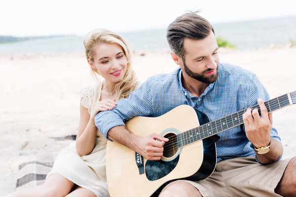 young blonde barefoot woman sitting on blanket near boyfriend with acoustic guitar at beach near sea