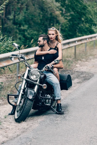 緑の森の近くの道路で黒いオートバイに抱きつく残忍なバイカーの若いカップル — ストック写真