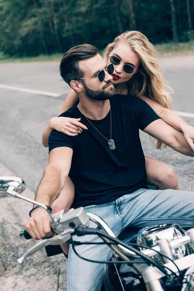 緑の森の近くの道路で黒いオートバイに抱きつくサングラスの若いカップル — ストック写真