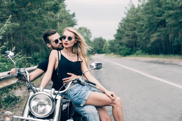 緑の森の近くの道路で黒いオートバイに密接に座っているバイカーの若いカップル — ストック写真