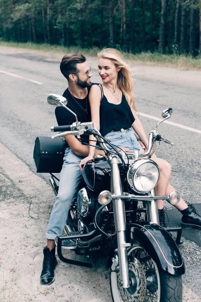 緑の森の近くの道路に黒いオートバイに座っているバイカーの幸せな若いカップル — ストック写真