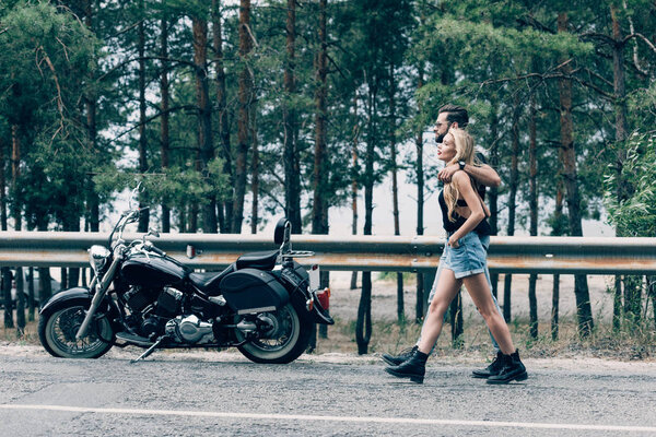 side view of young couple of bikers walking along road and embracing near black motorcycle and green forest