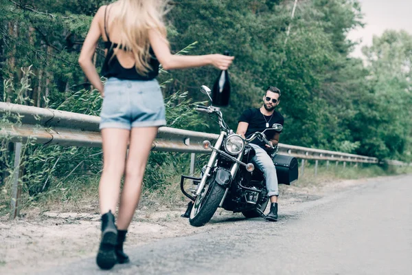 道路上の黒いオートバイのボーイフレンドの近くにアルコールのボトルを持って道路を歩いている若い女性のバックビュー — ストック写真