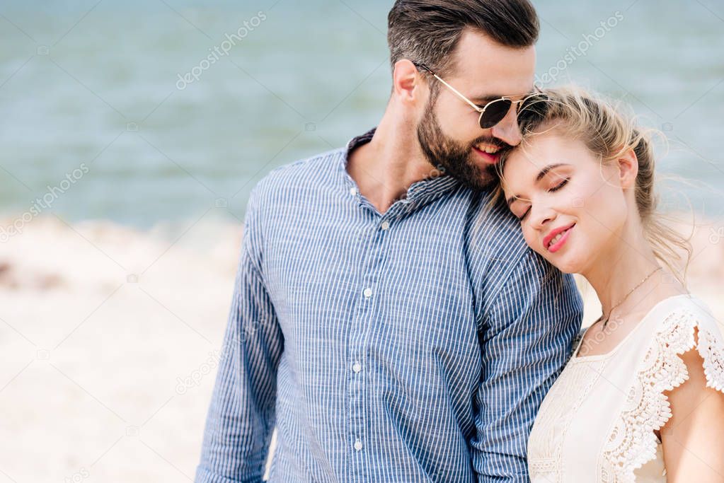 blonde girl with closed eyes leaning on boyfriend shoulder while man smiling at beach