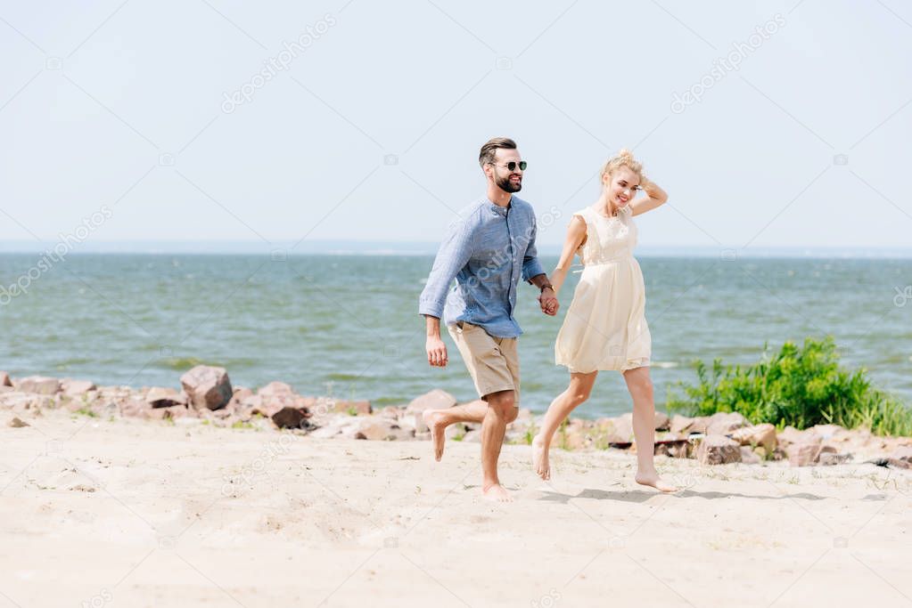 smiling young couple holding hands while running along beach