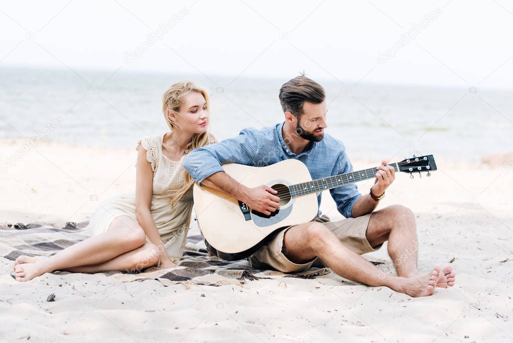 beautiful young barefoot woman sitting on blanket with boyfriend playing acoustic guitar at beach near sea